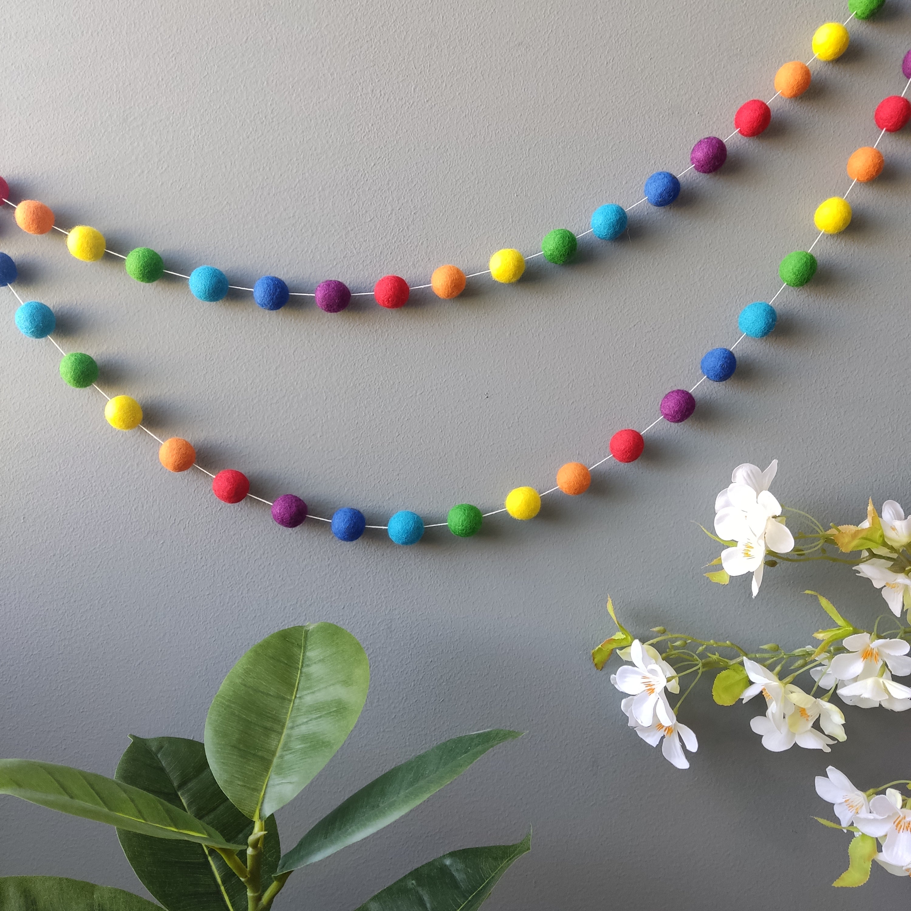 Guirlande de pompons arc en ciel vue de loin en double accrochage sur un mur avec des fleurs en decoration