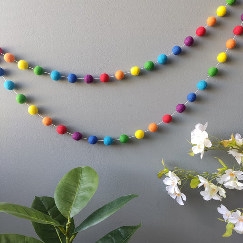 Guirlande de pompons arc en ciel vue de loin en double accrochage sur un mur avec des fleurs en decoration