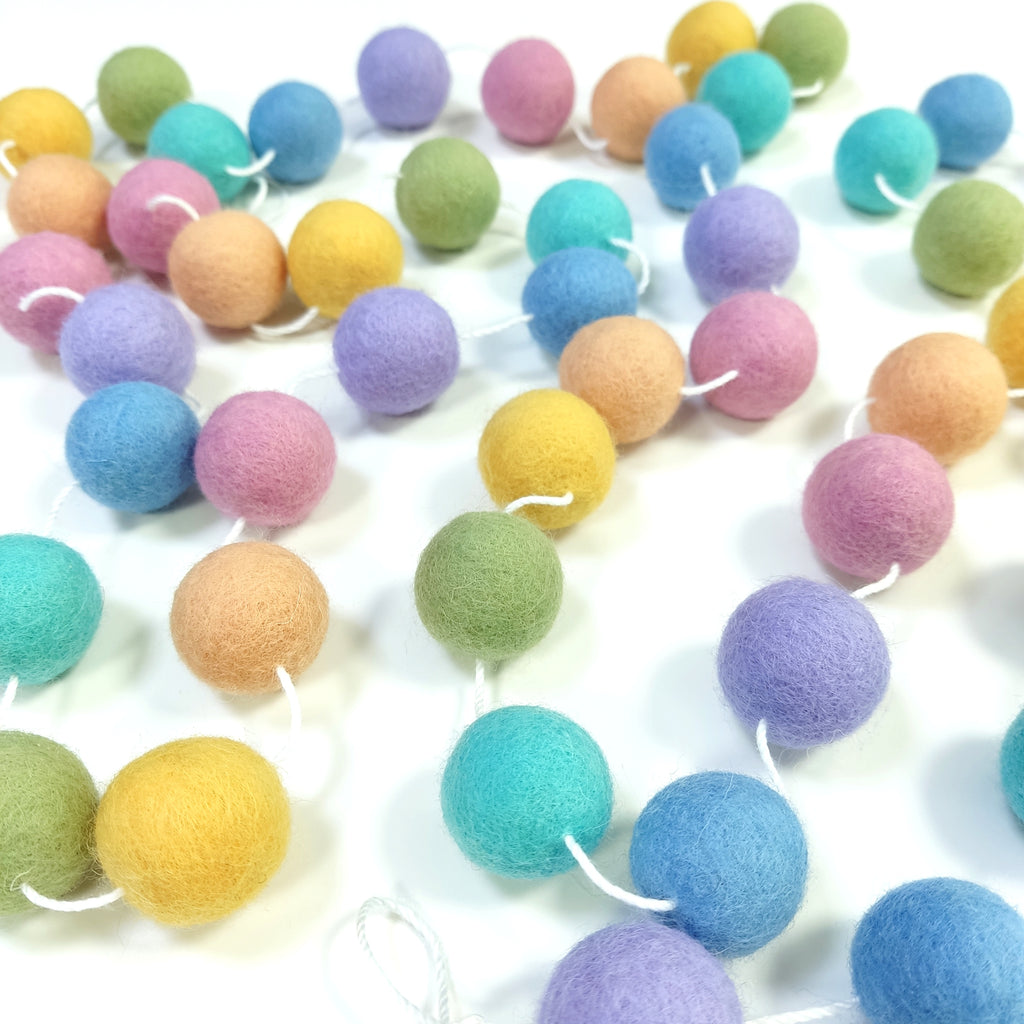 Guirlande en pompons de feutre pastel étalée sur une une table avec les pompons espacés