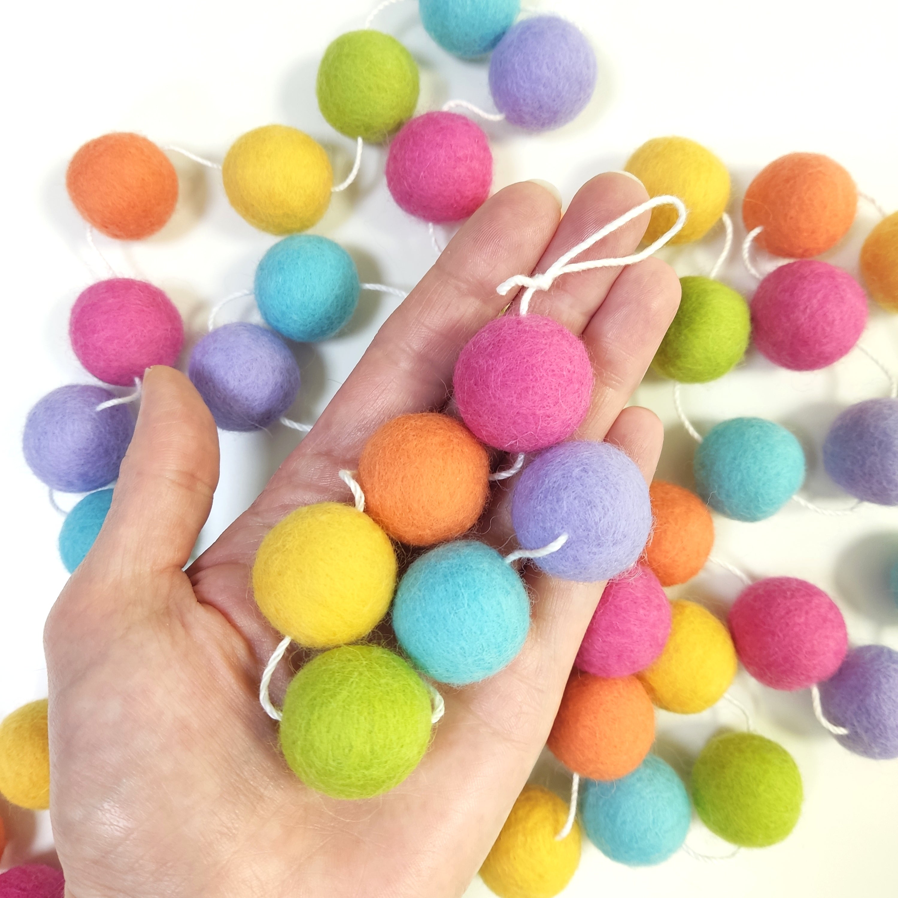 Détails des pompons de la guirlande rose bonbon, orange flamingo, jaune maïs, vert citron, bleu aqua et lavande dans une main