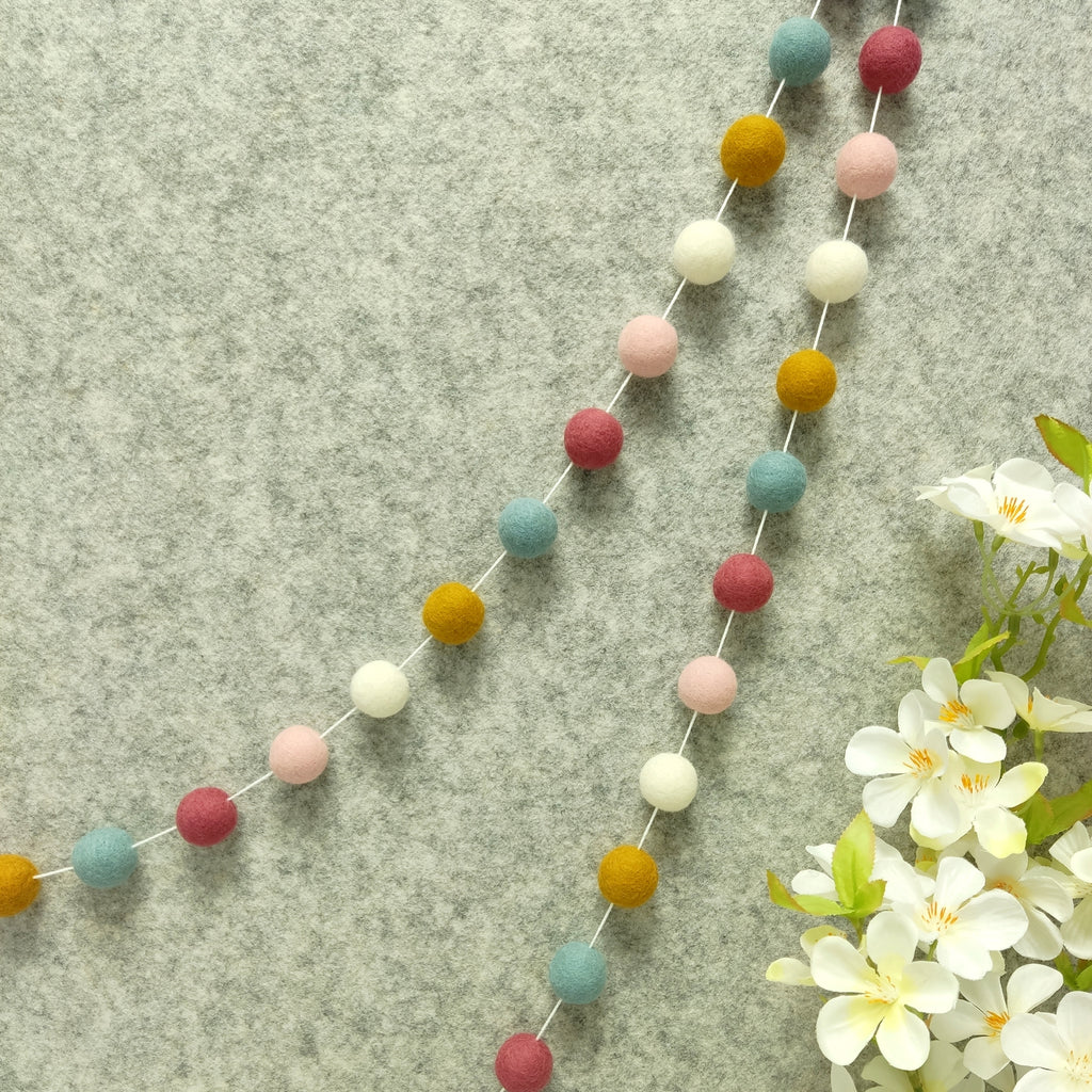 Guirlande de pompons feutrés aube scandinave suspendue sur un mur, teintes blanc, ocre doré, bleu nordique, rose poudré et rose boisé