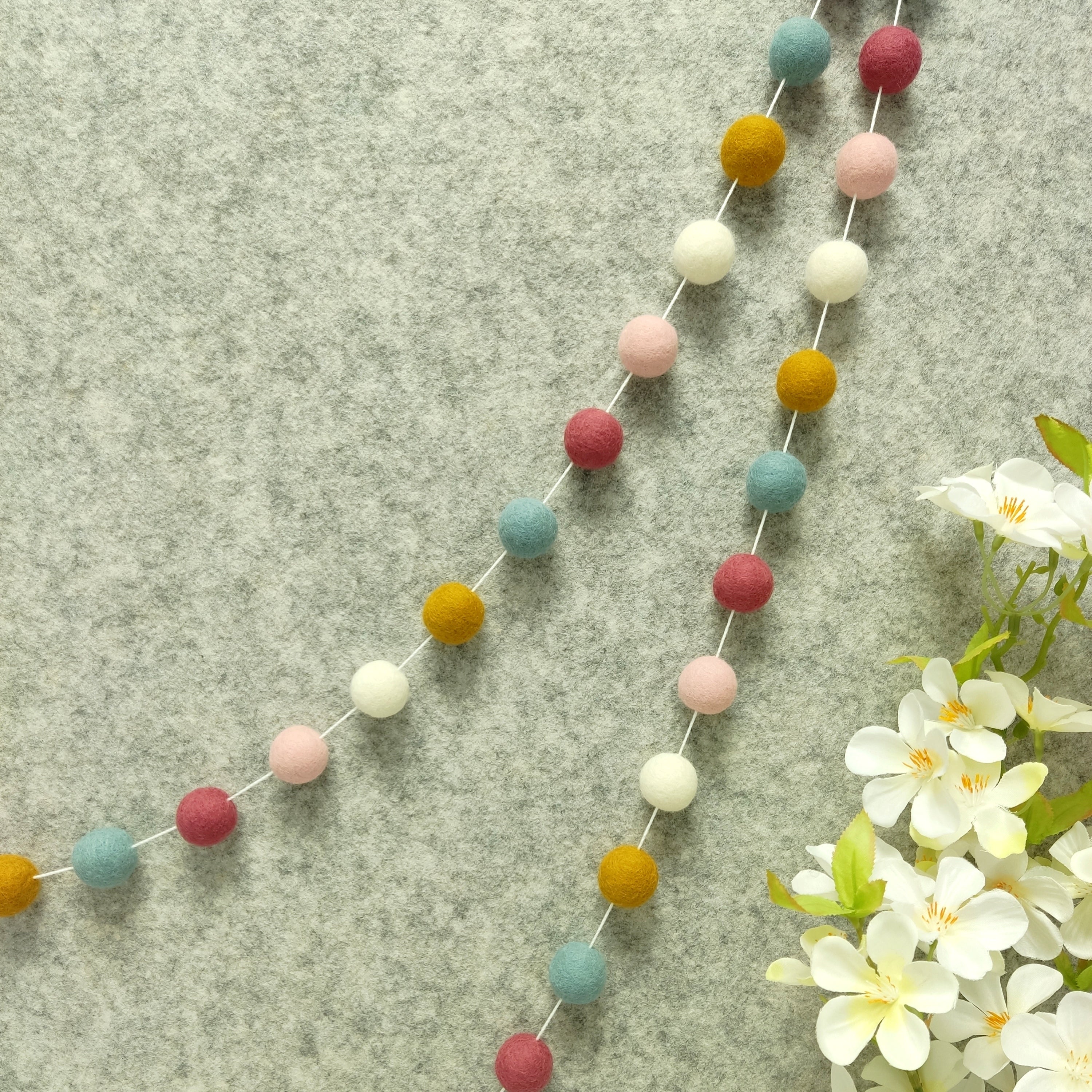 Guirlande de pompons feutrés aube scandinave suspendue sur un mur, teintes blanc, ocre doré, bleu nordique, rose poudré et rose boisé