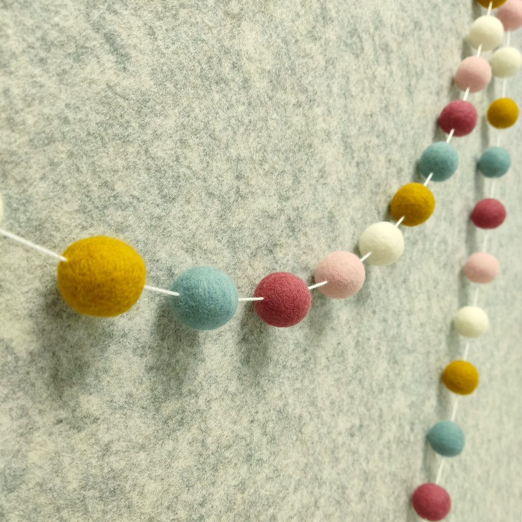 Guirlande de pompons feutrés aux teintes nordiques blanc, ocre, bleu nordique et rose poudré vue de profil