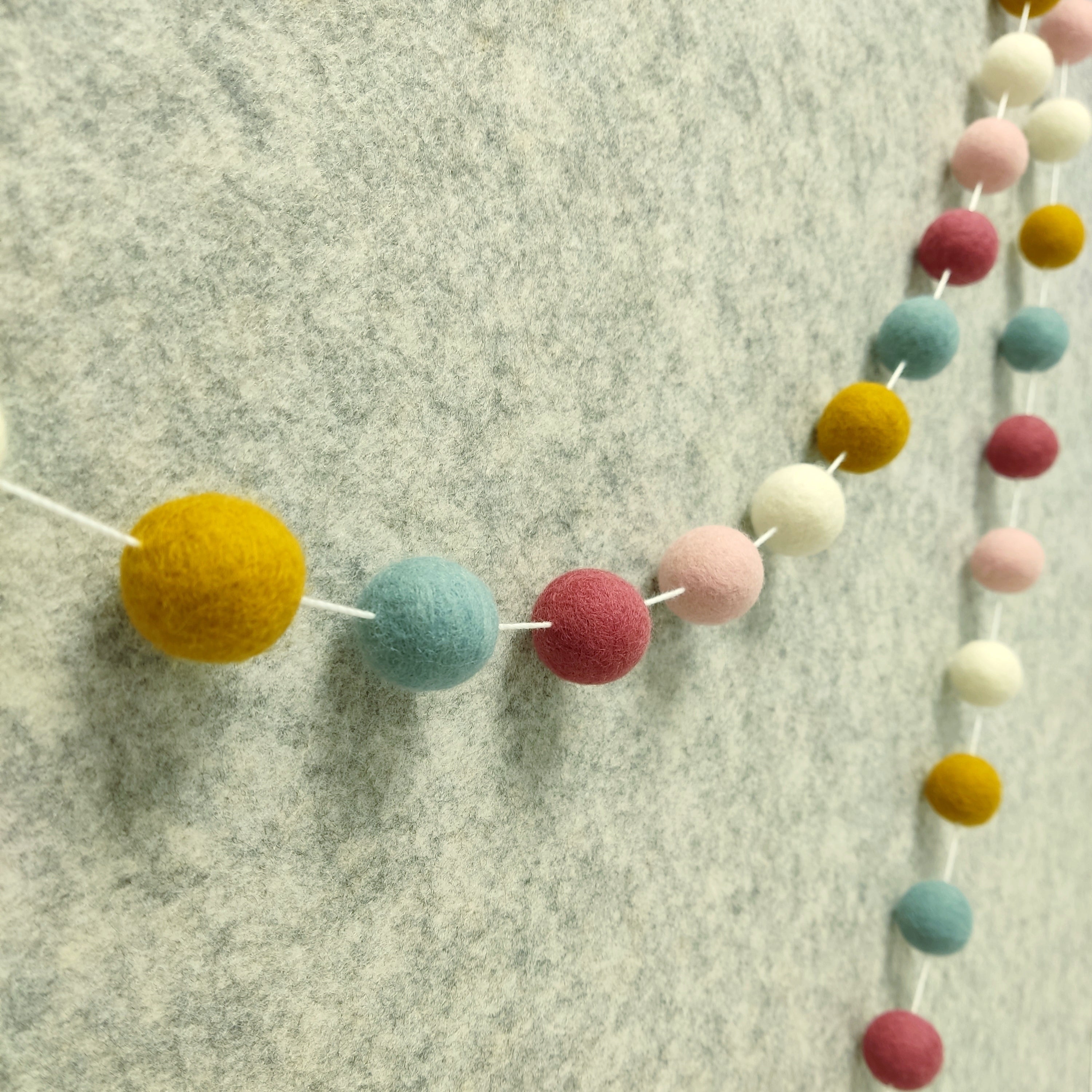 Guirlande de pompons feutrés aux teintes nordiques blanc, ocre, bleu nordique et rose poudré vue de profil