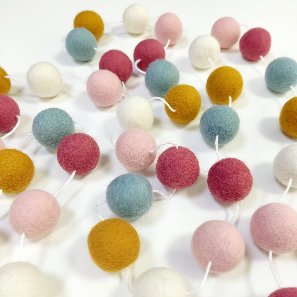 Guirlande de pompons feutrés aube scandinave étalée sur une table avec les pompons espacés sur le fil
