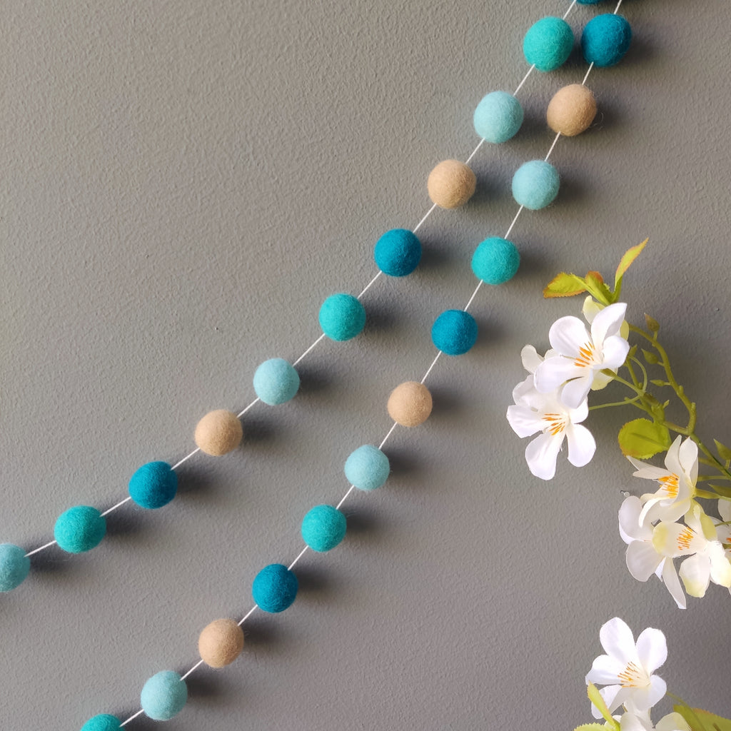 Guirlande de pompons feutrés beige et bleu turquoise suspendue sur un mur vue de près