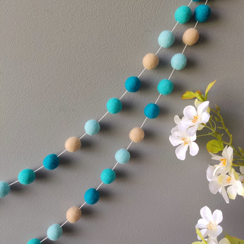 Guirlande de pompons feutrés beige et bleu turquoise suspendue sur un mur vue de près