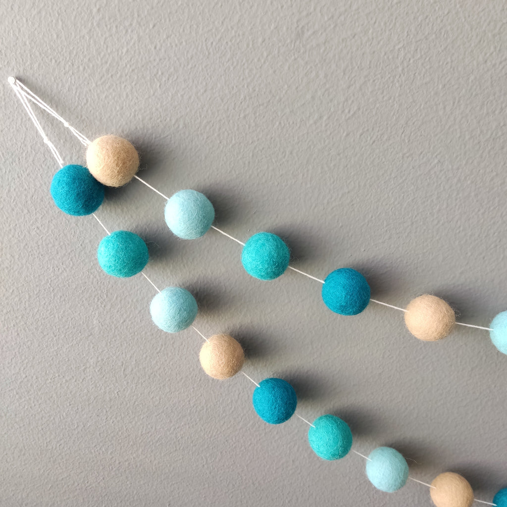 Détails des pompons en feutre beige et turquoise de la guirlande fixée au mur avec ses boucles aux extrémités