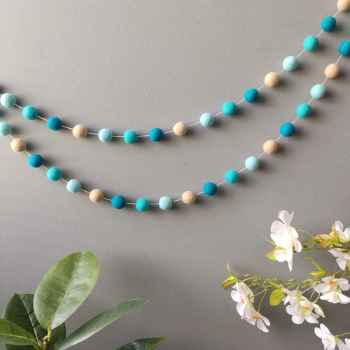 Guirlande de boules de feutre bleu turquoise et beige suspendue sur un mur en arc de cercle vue de loin