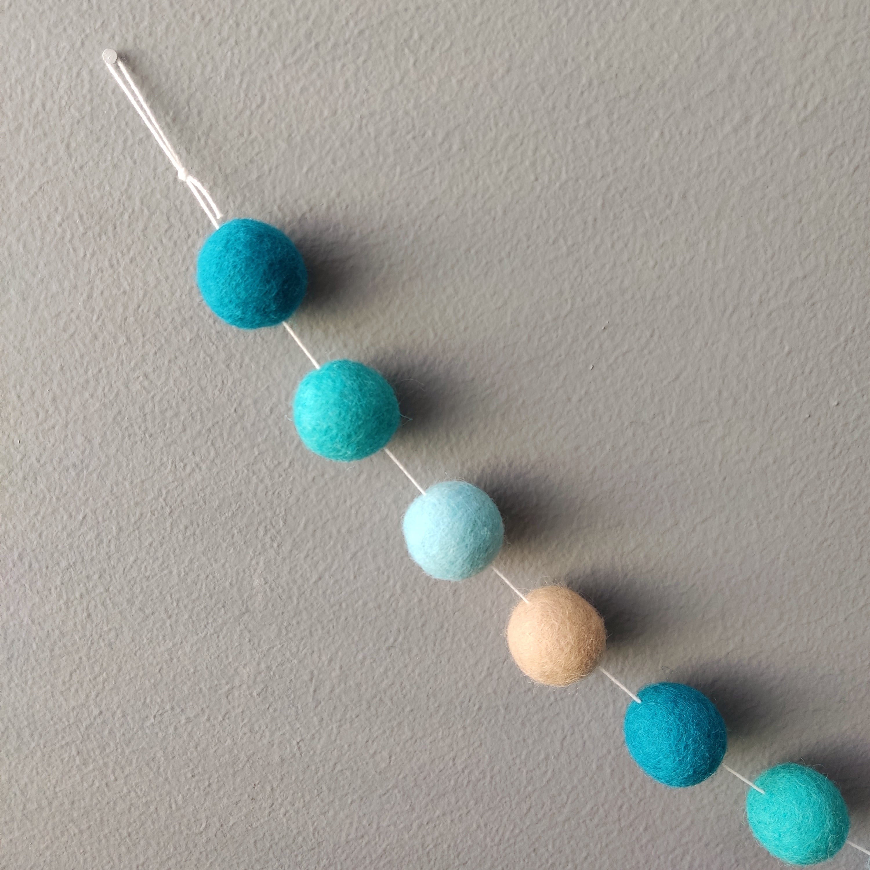 Zoom sur les pompons feutrés bleu turquoise, bleu canard, beige poudré et bleu aqua de la guirlande