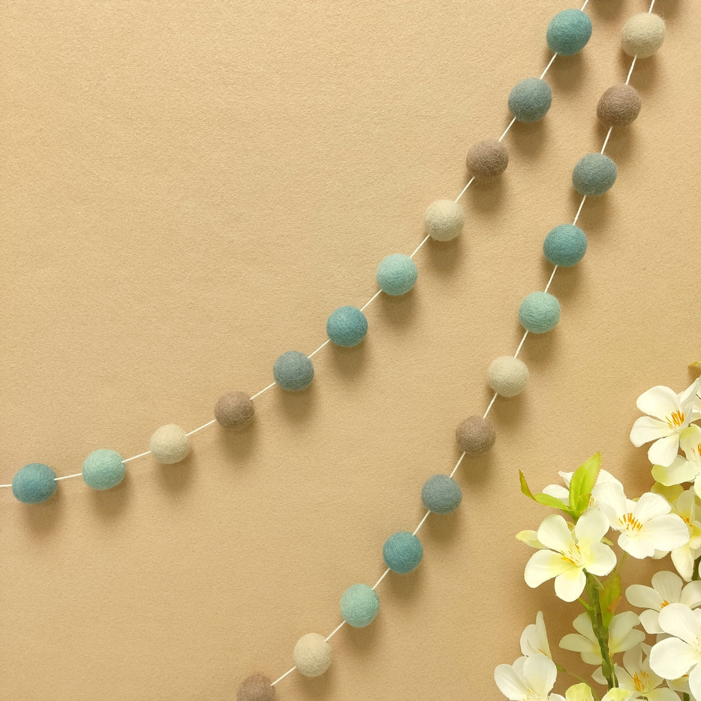 Guirlande pompons feutrés bleu beige gris vue de près suspendue sur un mur
