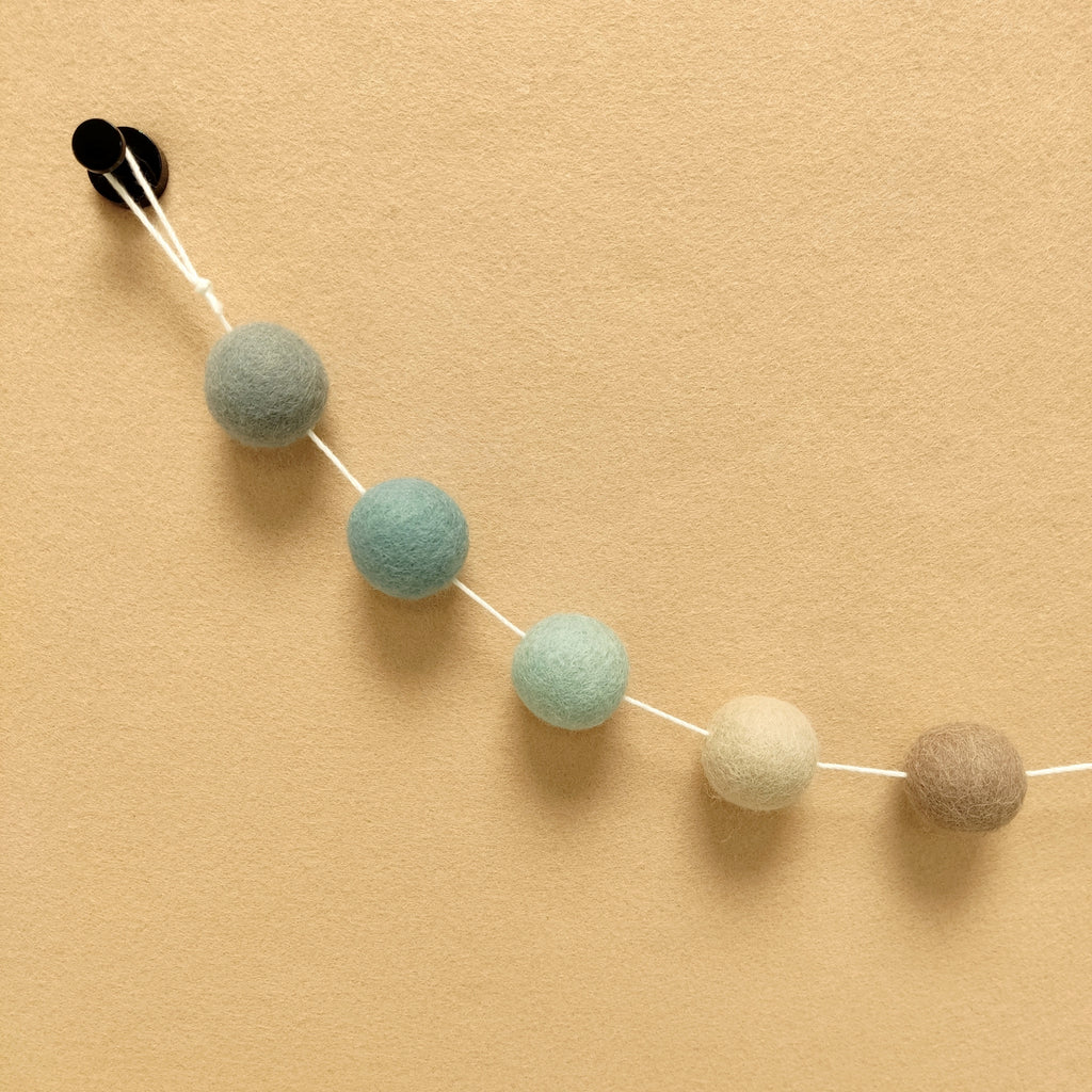 Zoom sur les pompons beige argile, vert céladon, bleu nordique, beige cappuccino et gris galet de la guirlande avec la boucle de fixation