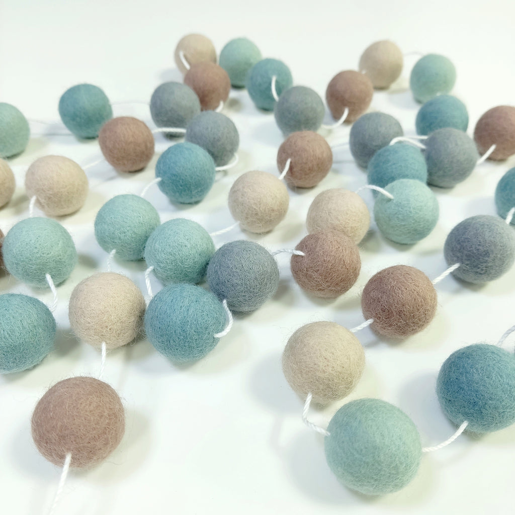 Guirlande de boules de laine feutrée bleu beige gris vert étalée sur une table