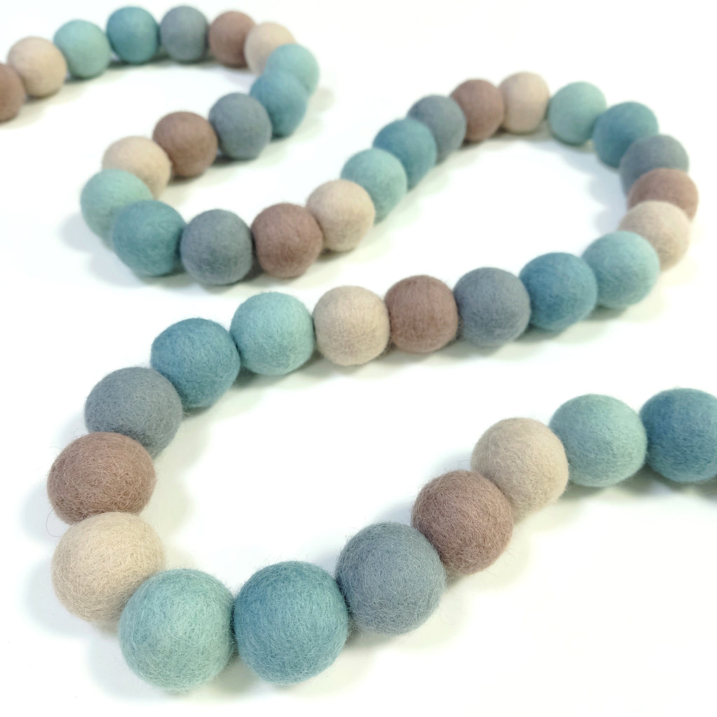 Guirlande de pompons feutrés bleu vert beige gris disposée en serpentin sur une table