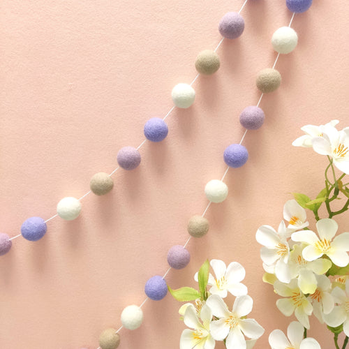 Guirlande de pompons feutrés blanc, beige, lilas et lavande fixée sur un mur