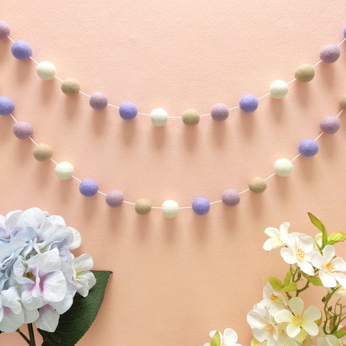 Guirlande en boules de feutre aux teintes beige, blanc mauve suspendue en arc de cercle sur un mur