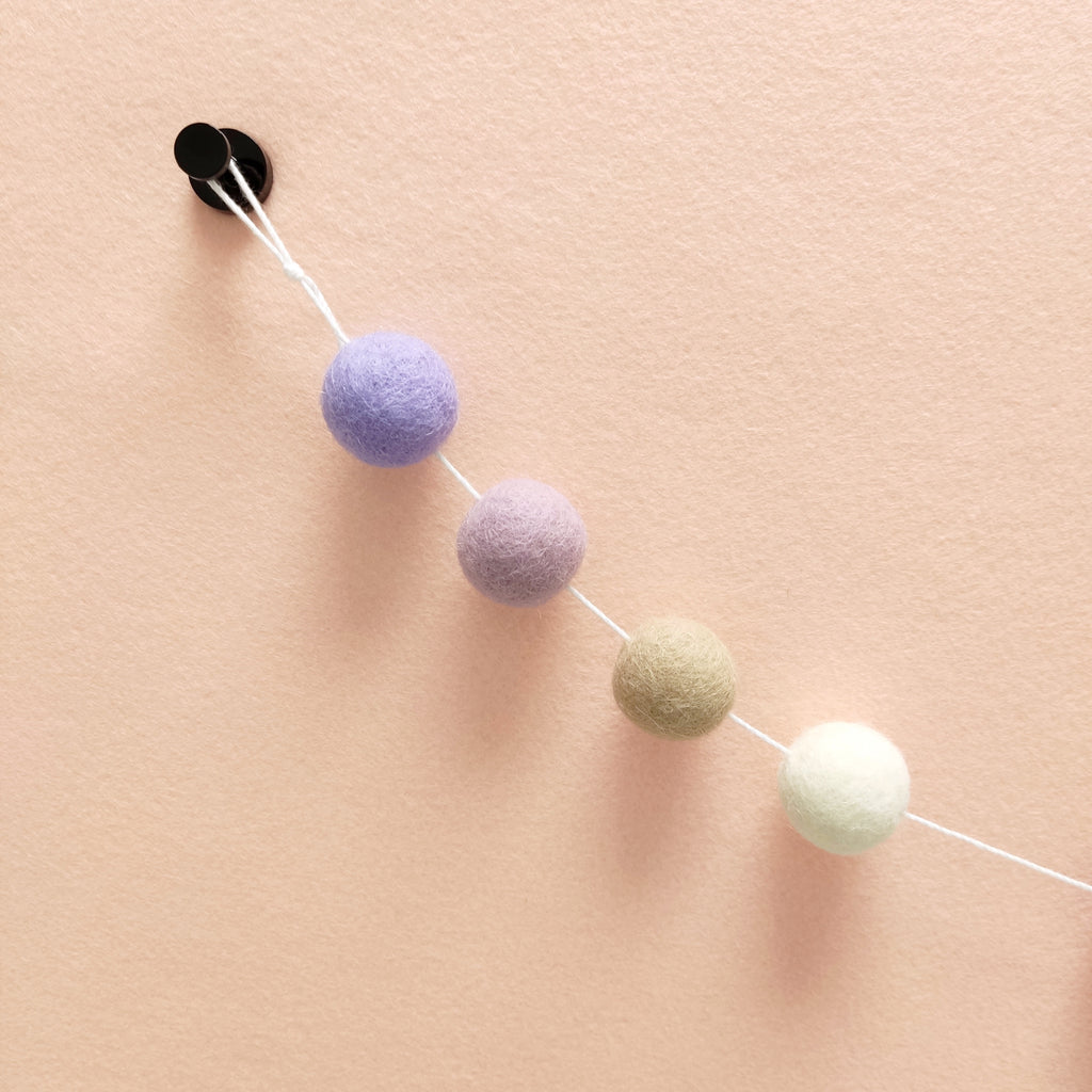 Détail des pompons en feutre de laine mérinos blanc naturel, beige poudré, lilas et lavande sur le fil de la guirlande avec la boucle de fixation