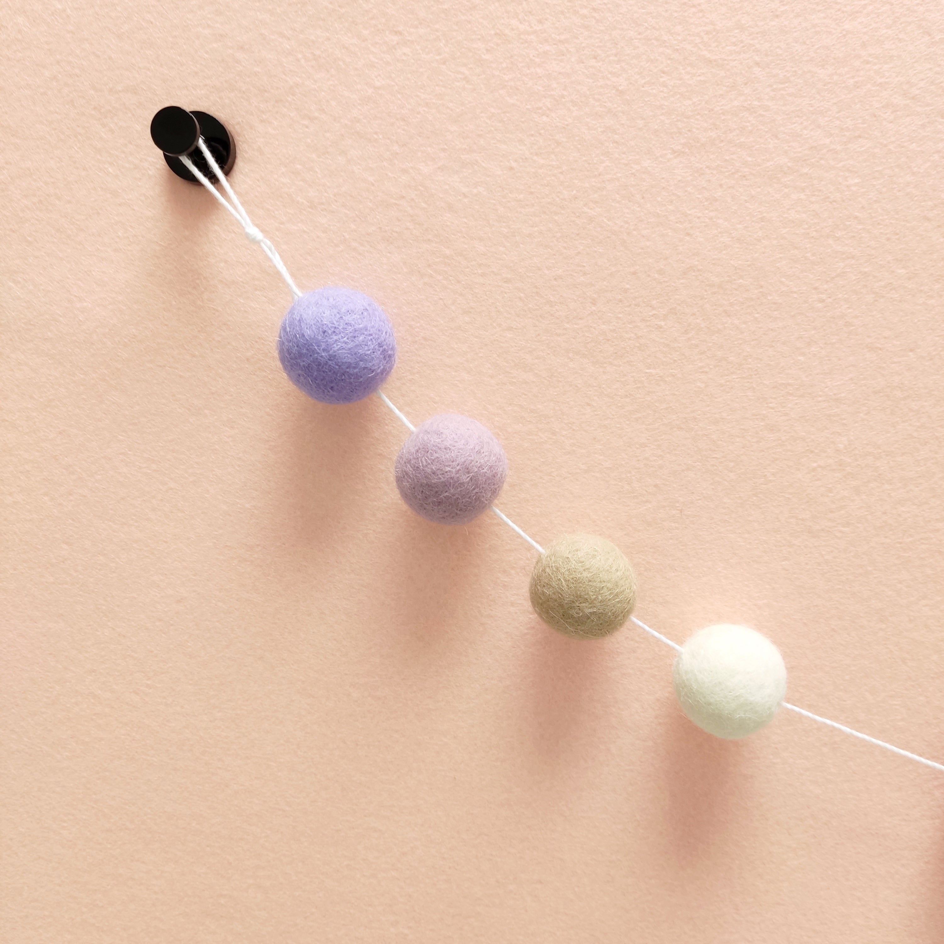 Détail des pompons en feutre de laine mérinos blanc naturel, beige poudré, lilas et lavande sur le fil de la guirlande avec la boucle de fixation