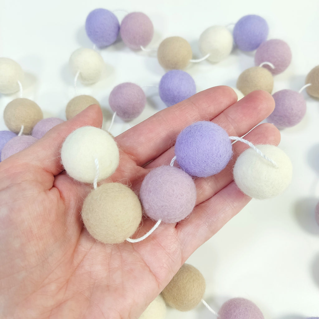 Zoom sur les pompons en feutre de laine mérinos blanc, beige poudré, lilas et lavande de la guirlande Brume Florale dans une main