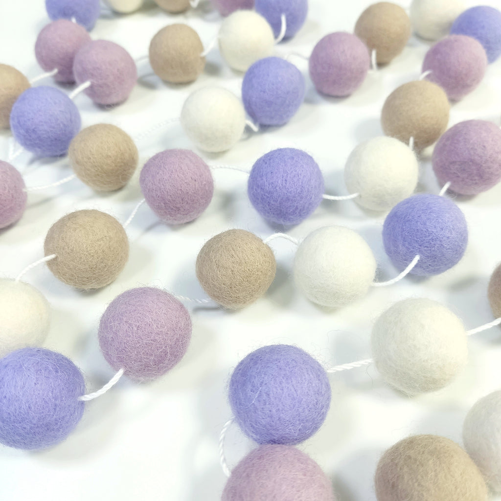 Guirlande de pompons feutrés blanc, beige, lilas et lavande étalée sur une table avec les pompons espacés