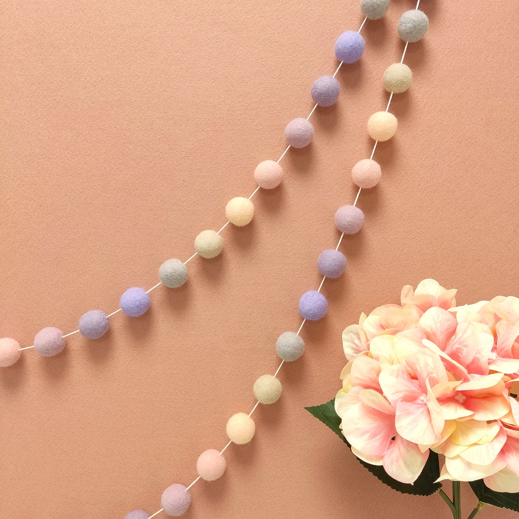Guirlande de pompons feutrés pastel beige rosé, rose poudré, lilas, mauve et lavande, suspendue sur un mur