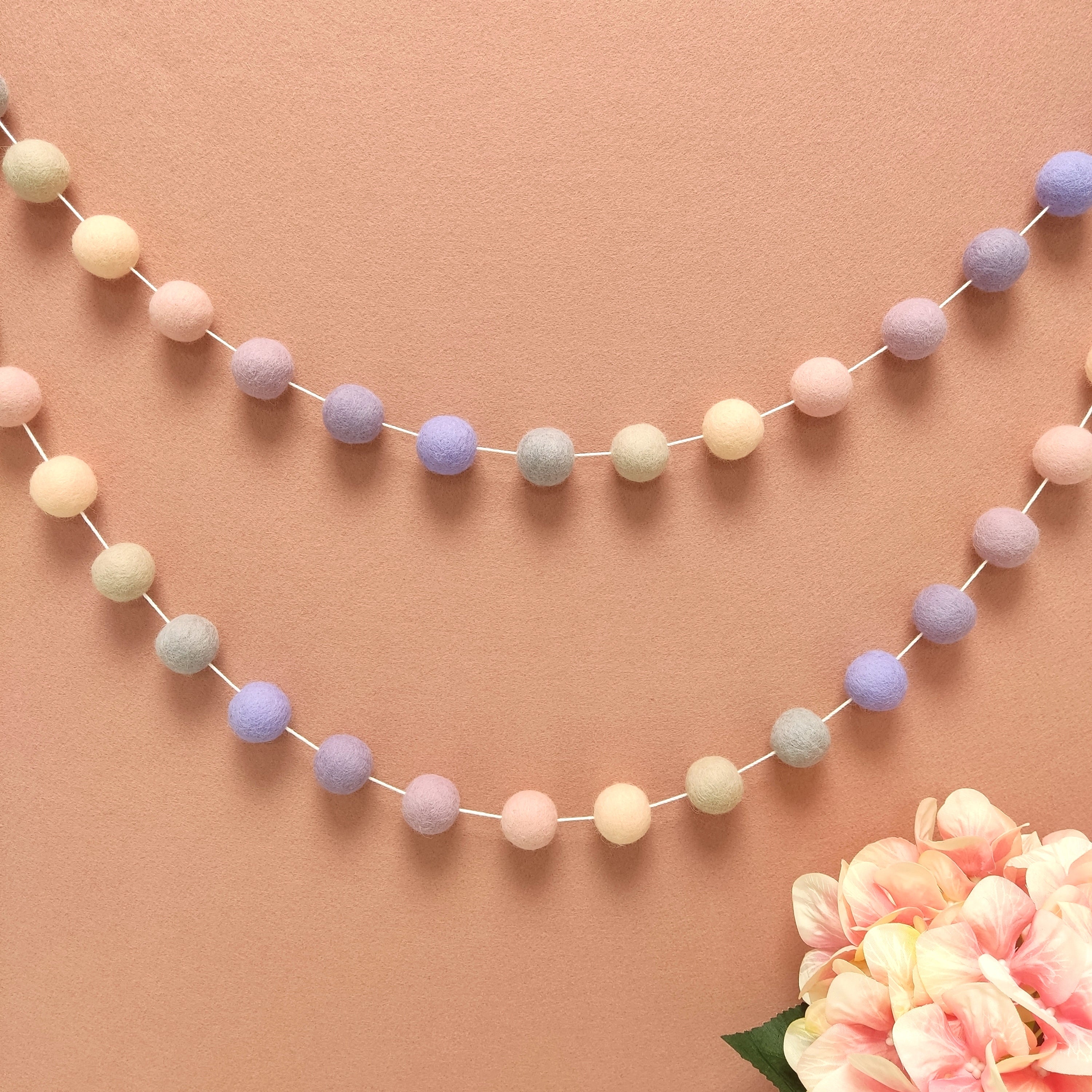 Guirlandes de pompons en feutre de laine mérinos, faite main en France, aux teintes beige, rose pastel et mauve, fixée en arc de cercle sur un mur