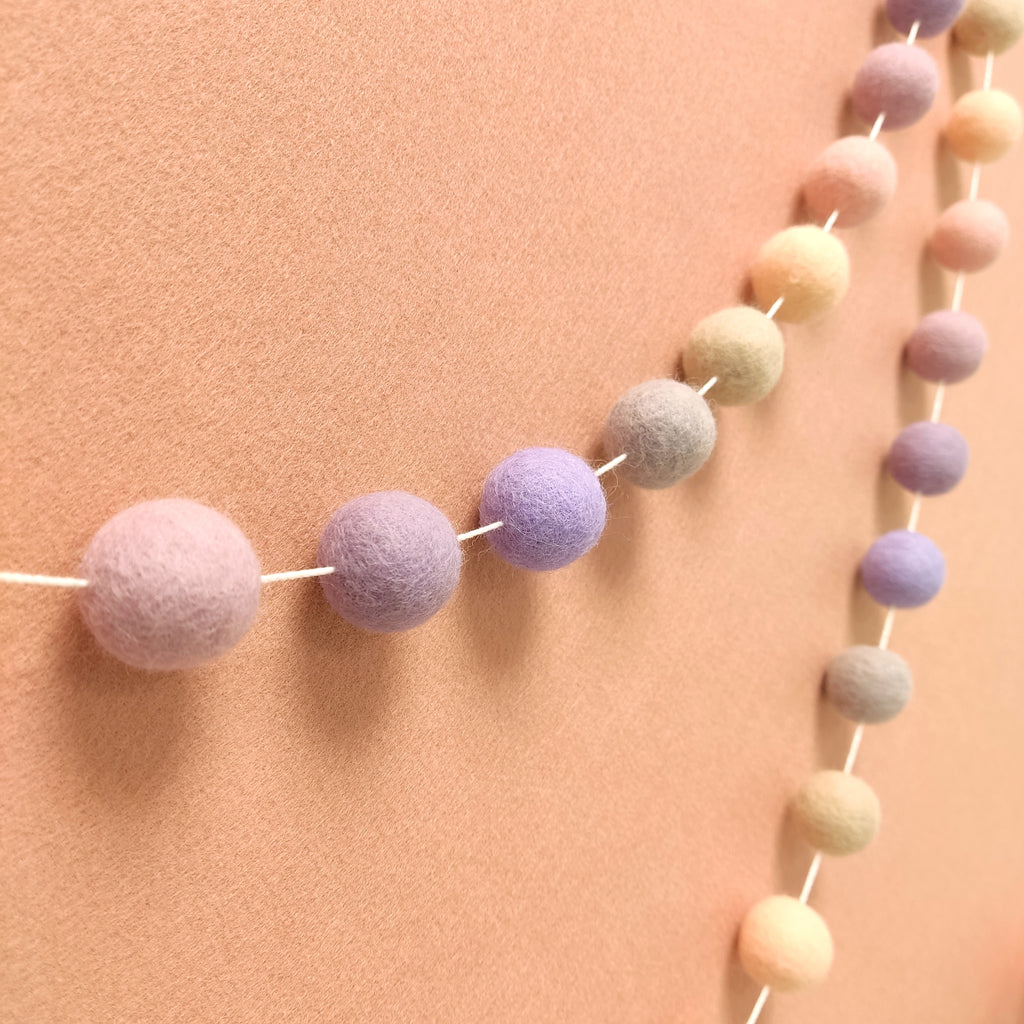 Guirlande de pompons en laine feutrée aux couleurs pastel rosé, beige, lilas et lavande, vue de profil sur un mur
