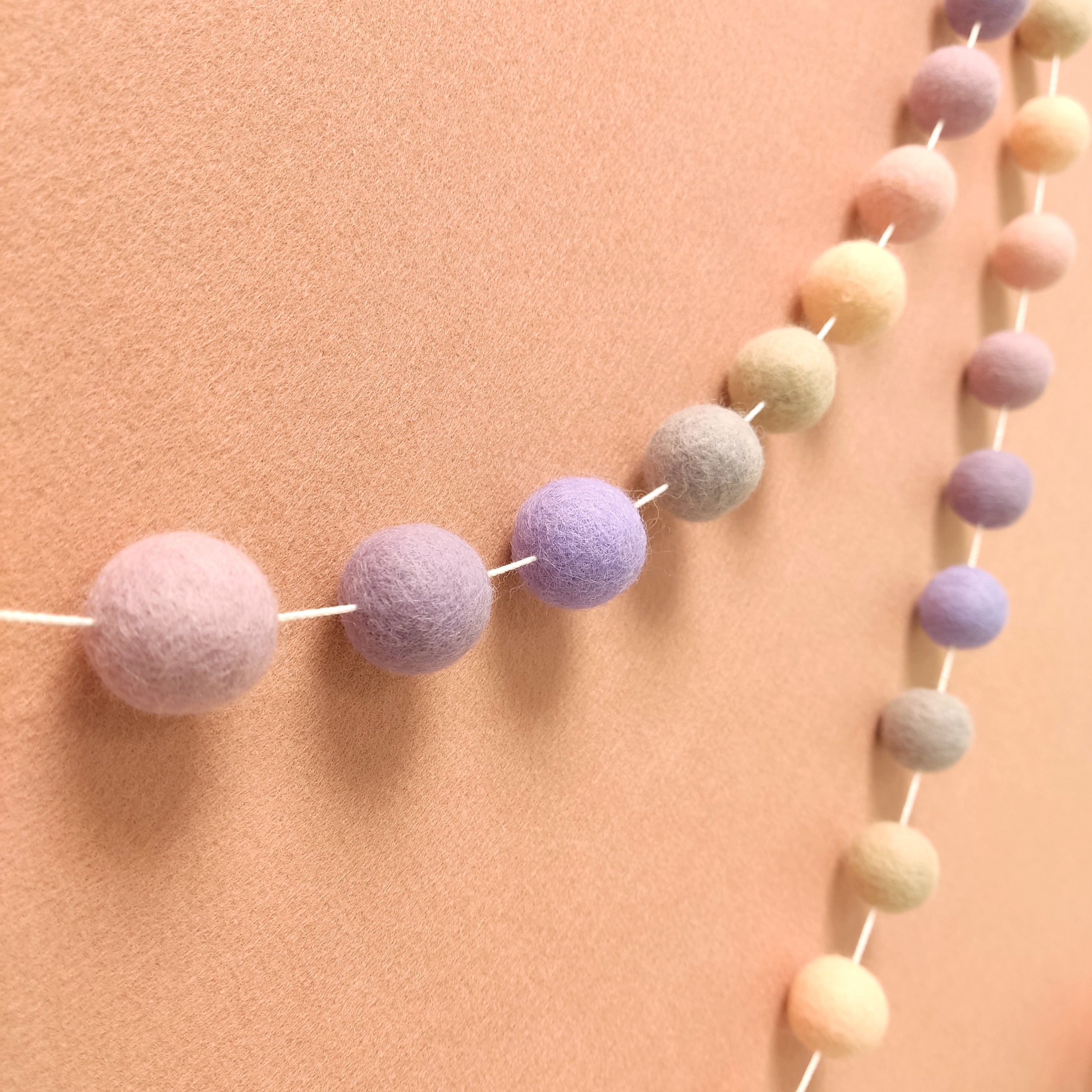 Guirlande de pompons en laine feutrée aux couleurs pastel rosé, beige, lilas et lavande, vue de profil sur un mur