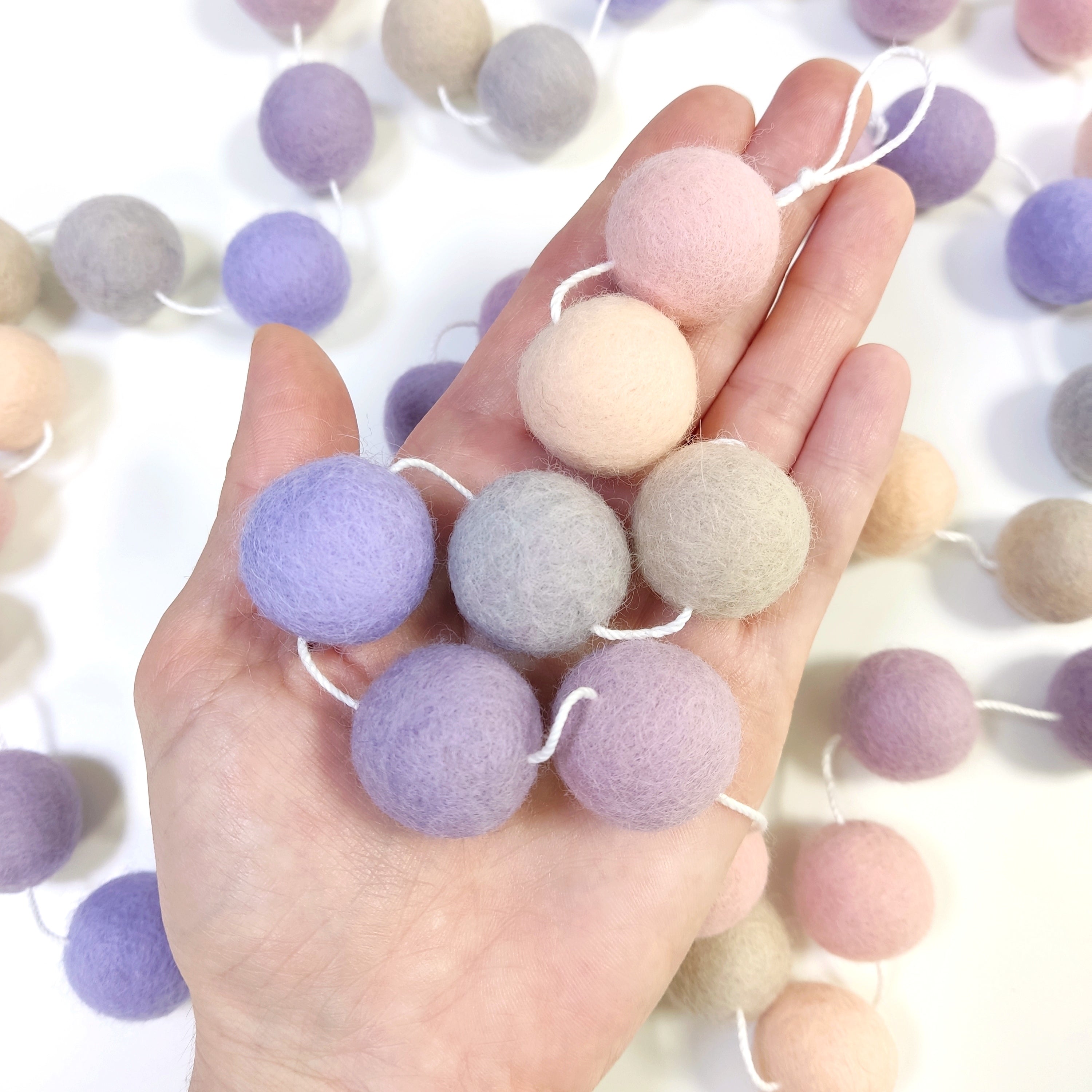 Zoom sur les pompons en laine feutrée de la guirlande beige, rose, mauve et lavande, dans une main