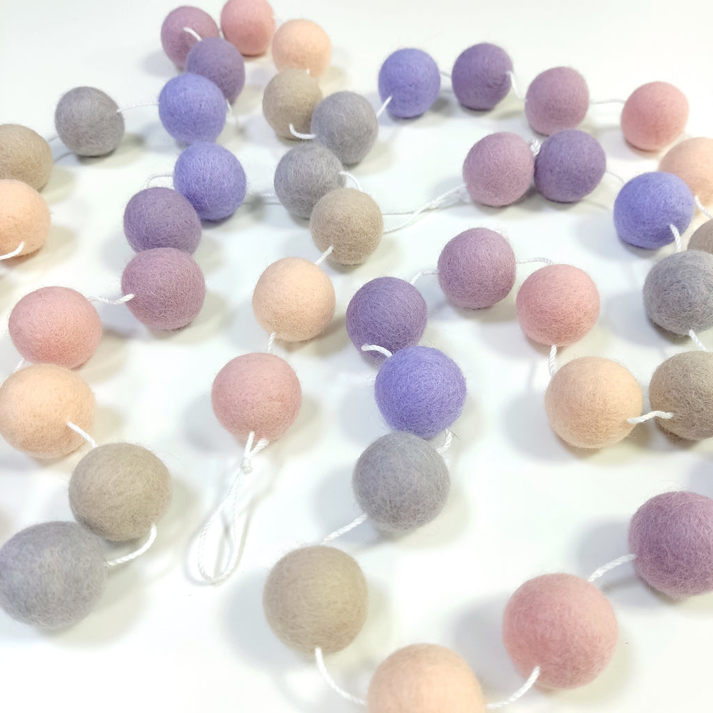 Guirlande en boules de feutre beige, rose et mauve, étalée sur une table, avec les pompons espacés