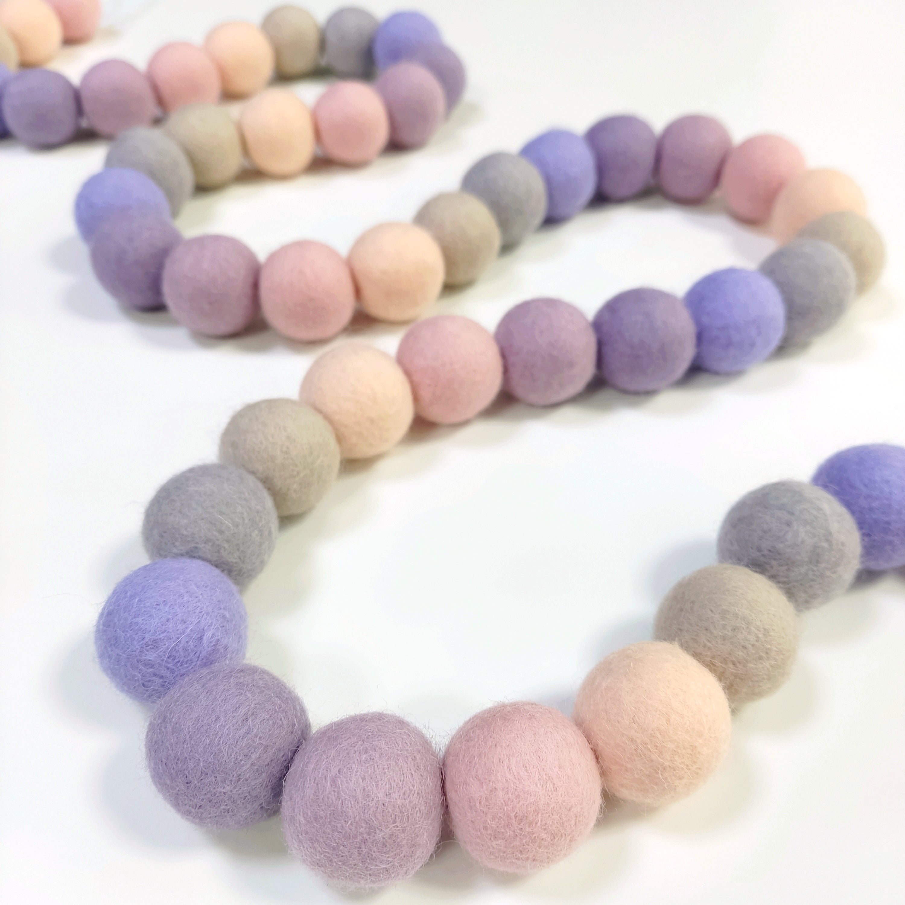 Guirlande de pompons feutrés aux tons pastels beige, rose, lilas et lavande, disposée en serpentin sur une table