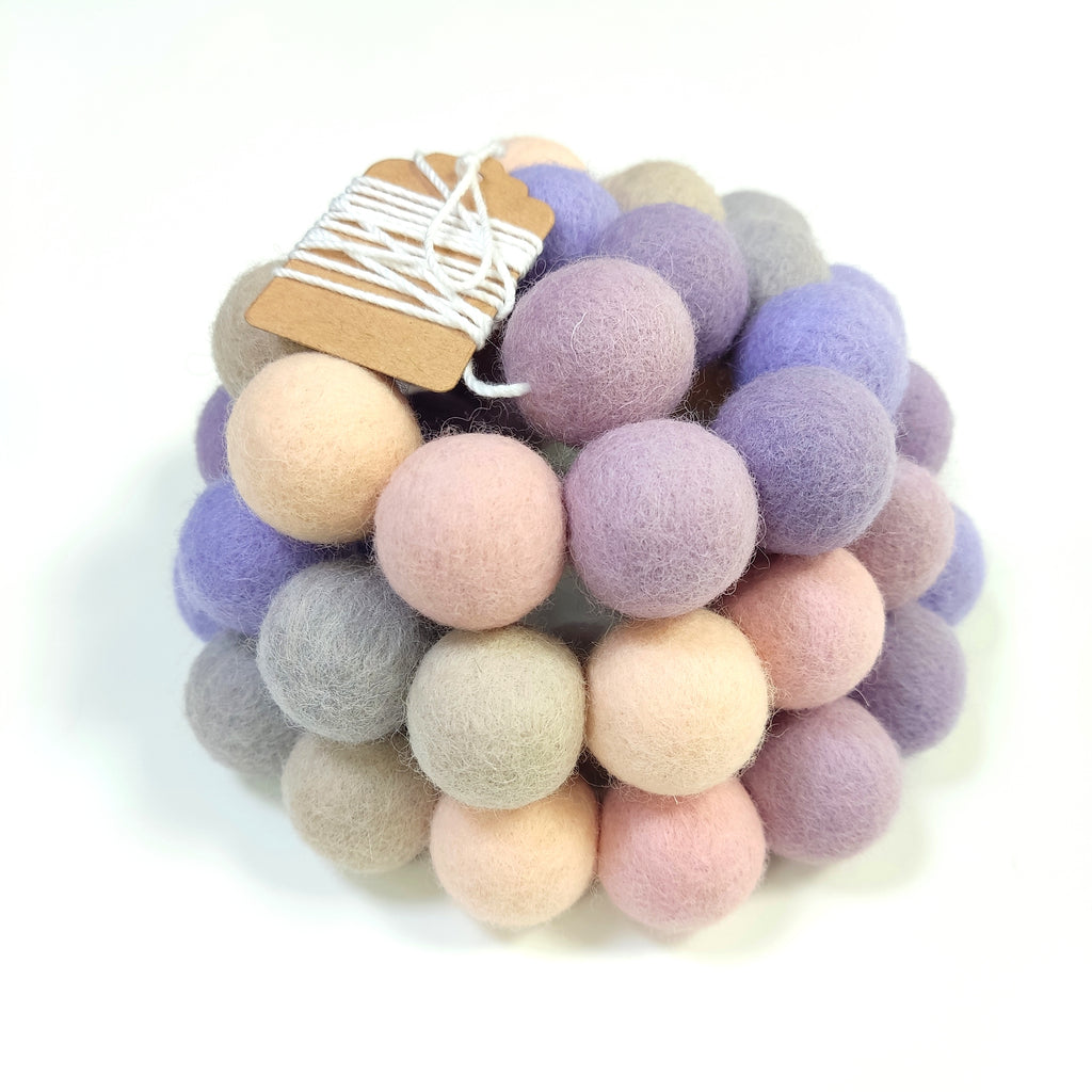 Zoom sur la guirlande en pompons de laine feutrée aux teintes lavande, beige et rose, avec l'étiquette