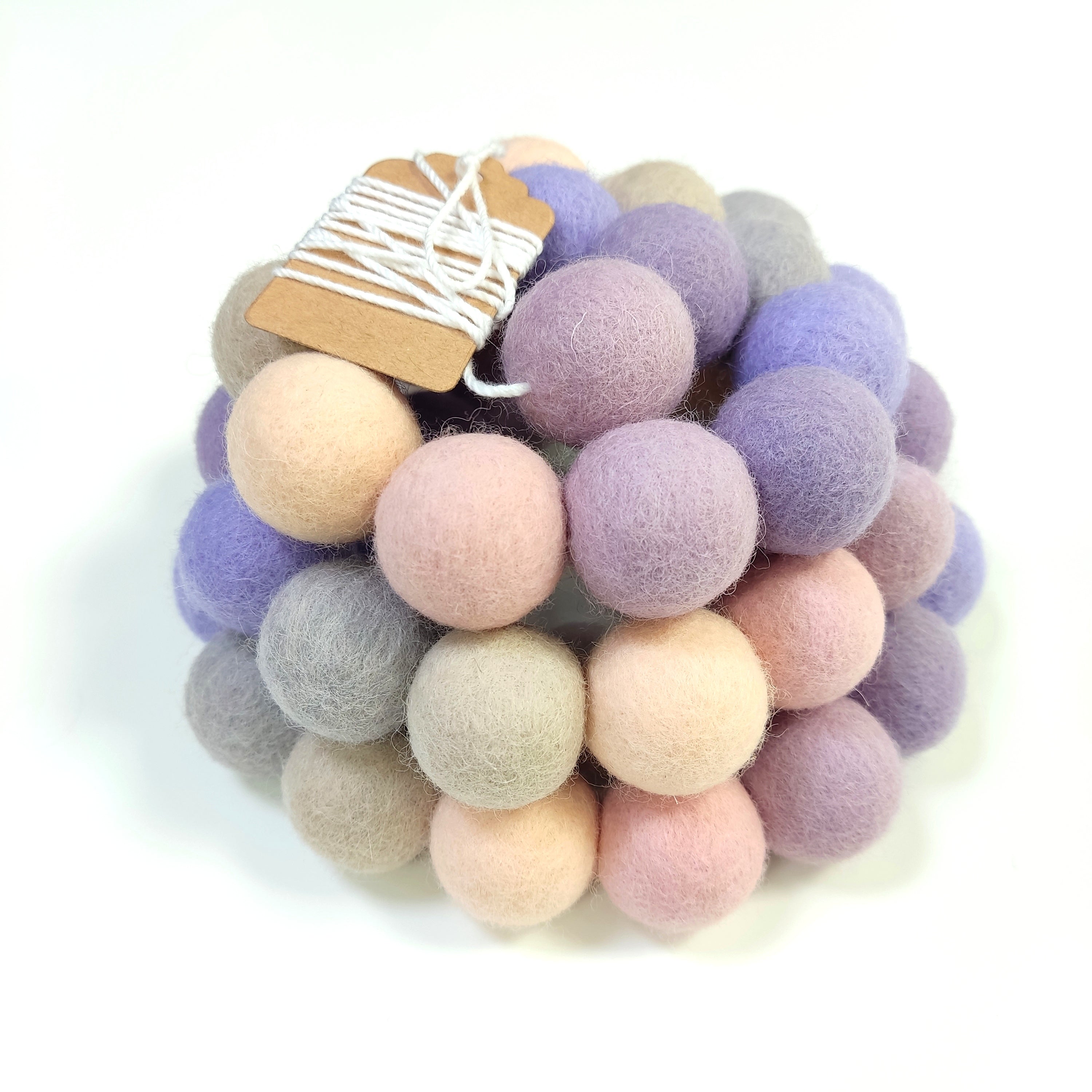 Zoom sur la guirlande en pompons de laine feutrée aux teintes lavande, beige et rose, avec l'étiquette