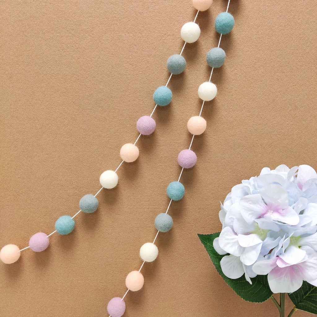 Guirlande de pompons feutrés gris, blanc, beige et bleu accrochée sur un mur de chambre