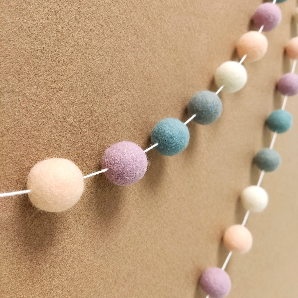 Guirlande de pompons en feutre de laine mérinos aux teintes blanc gris lilas vue de profil sur un mur