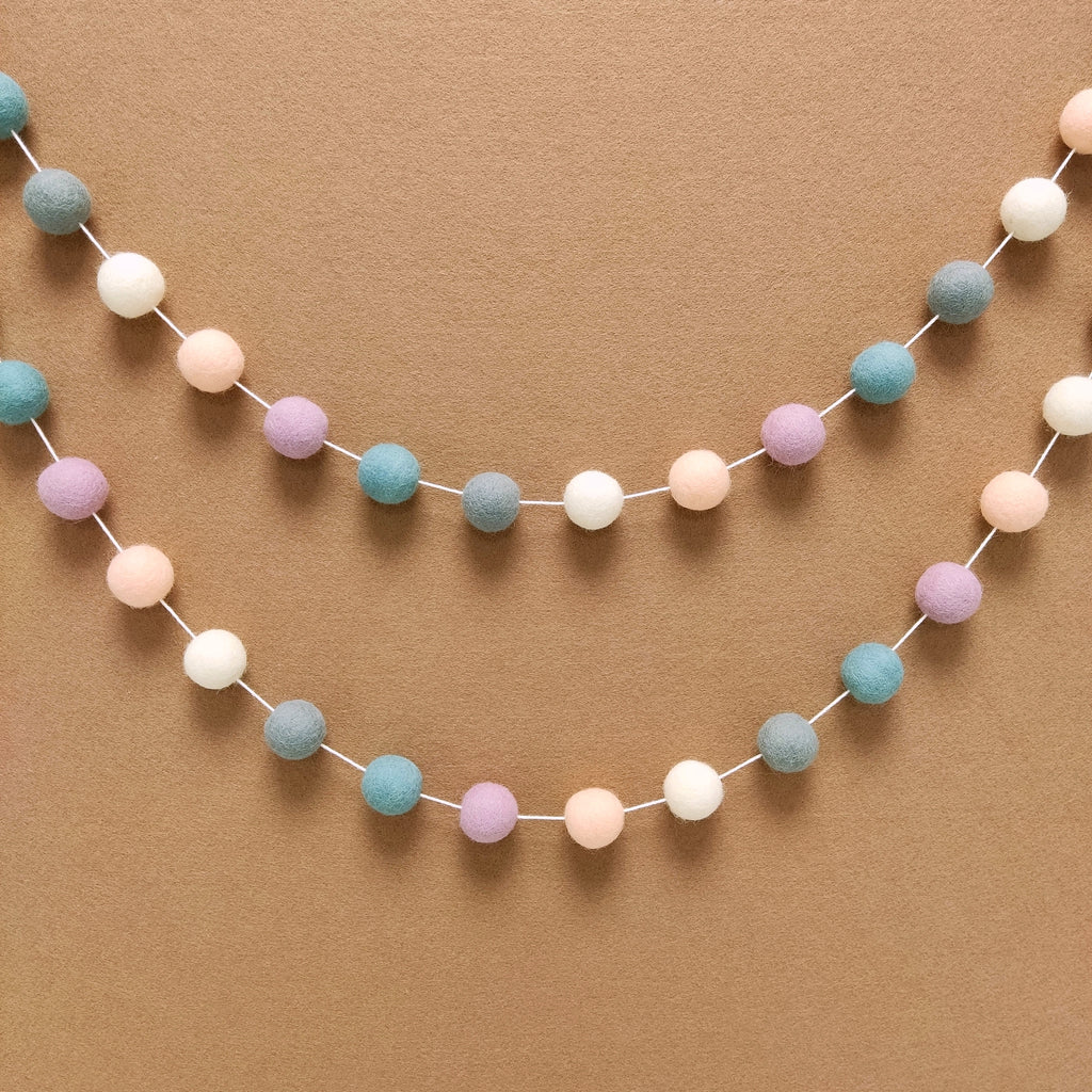 Guirlande en boules de feutre blanc, lilas, bleu nordique, beige rosé et gris galet, suspendue en arc de cercle sur un mur