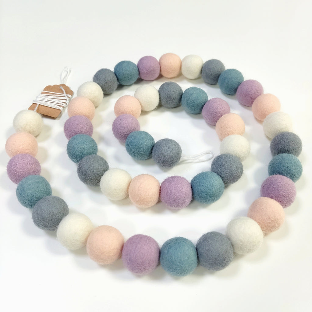 Guirlande de pompons beige rosé, blanc, gris, bleu et lilas, disposée en spirale sur une table