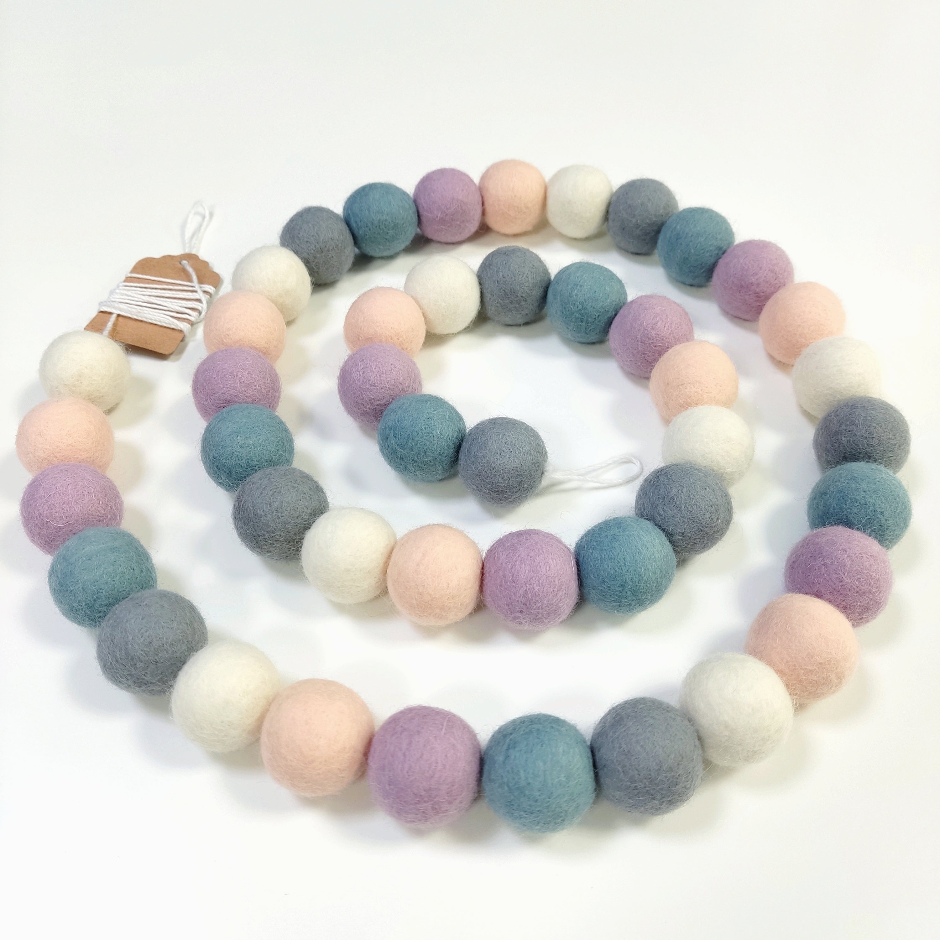 Guirlande de pompons beige rosé, blanc, gris, bleu et lilas, disposée en spirale sur une table