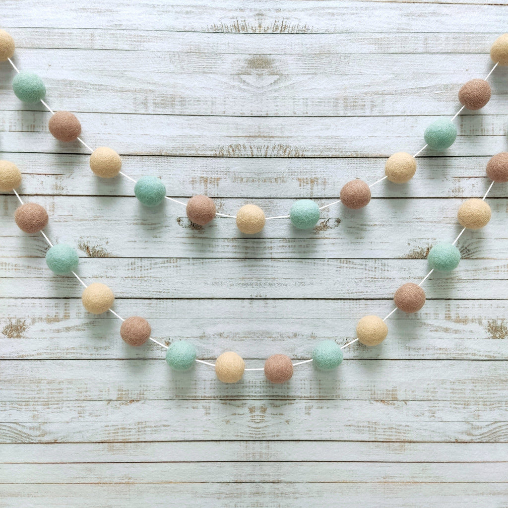 Guirlande de pompons vert céladon et beige fixée sur un mur en arc de cercle