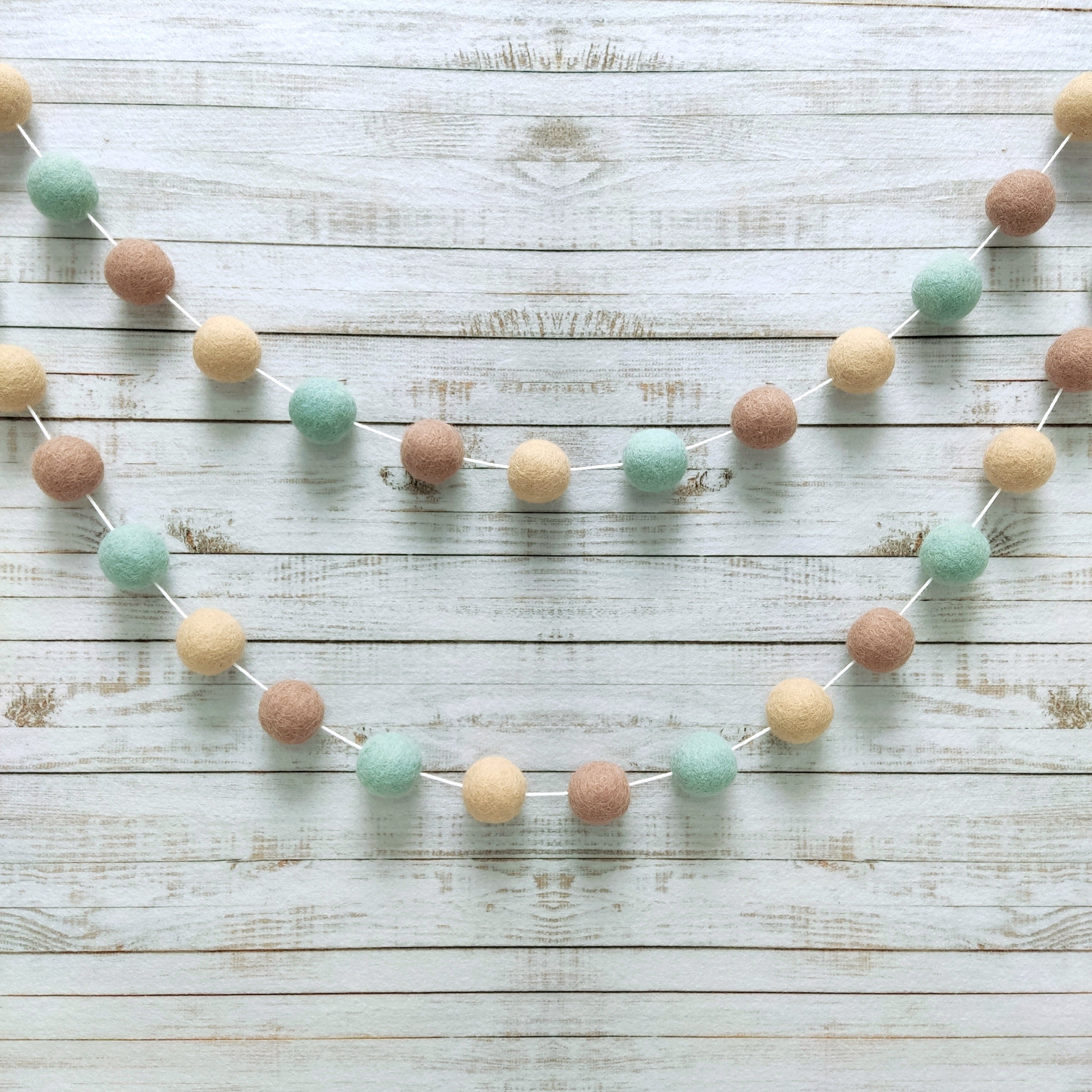 Guirlande de pompons vert céladon et beige fixée sur un mur en arc de cercle
