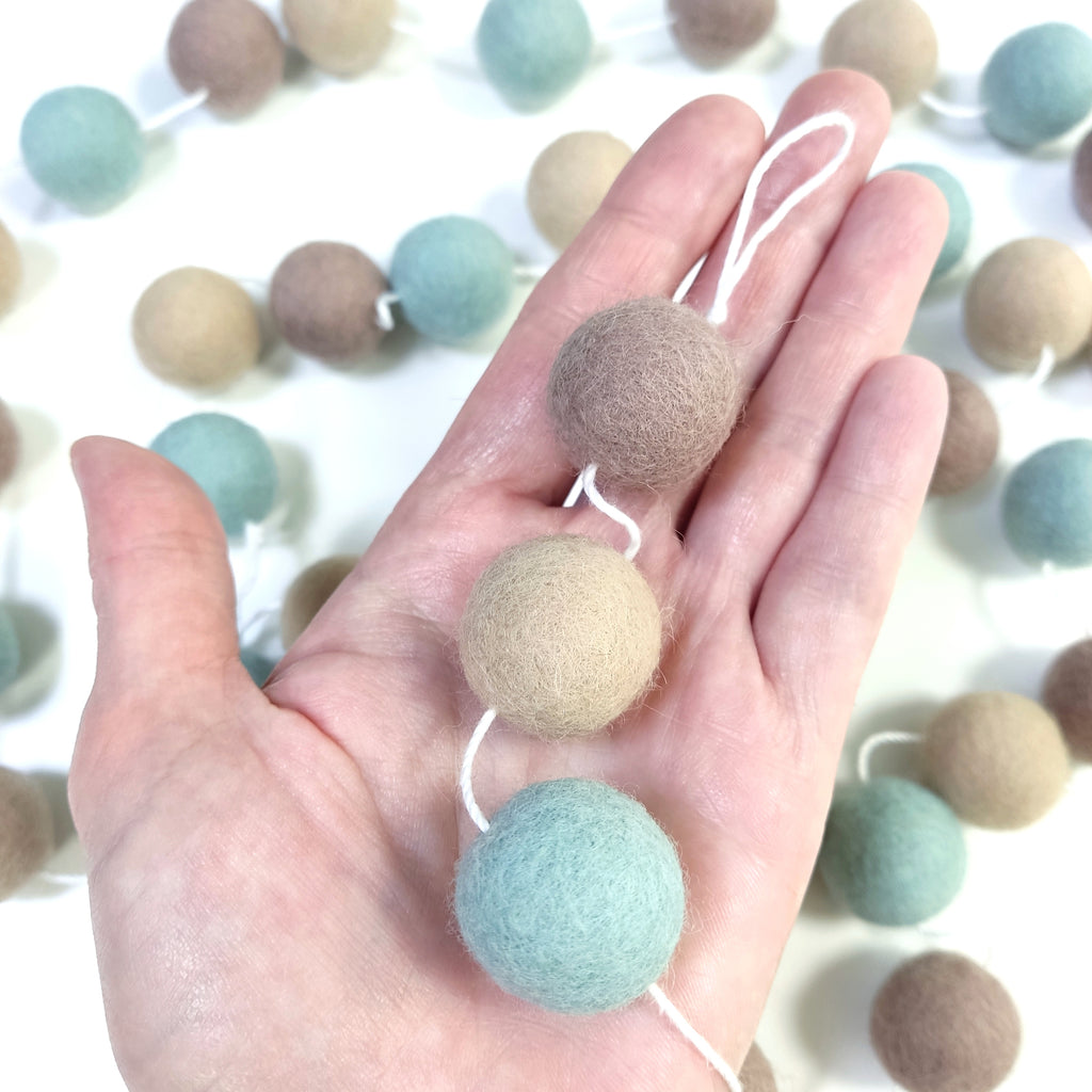 Pompons feutrés de la guirlande vert céladon, beige et cappuccino en détail dans une main