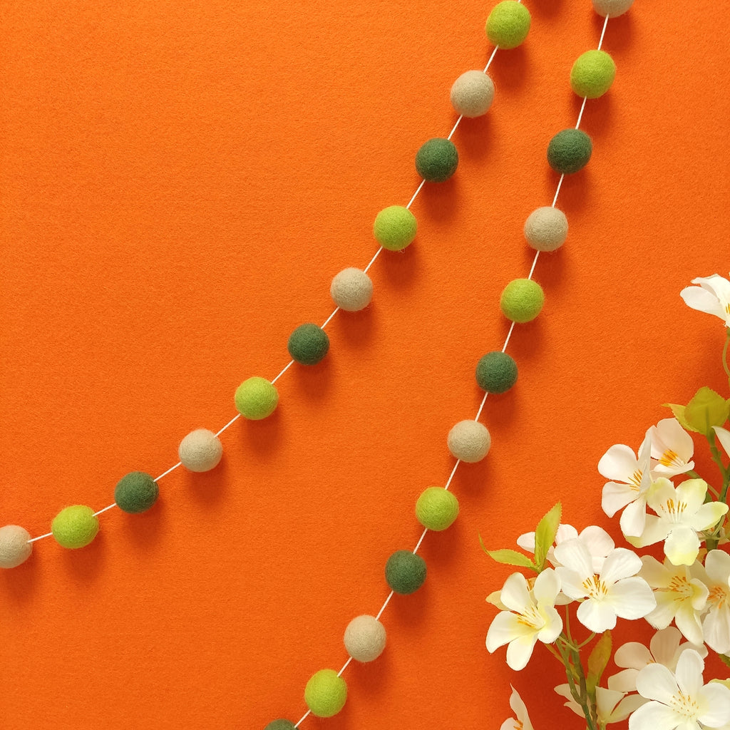 Guirlande de pompons en laine feutrée vert citron, vert matcha, beige poudré, accrochée sur un mur.