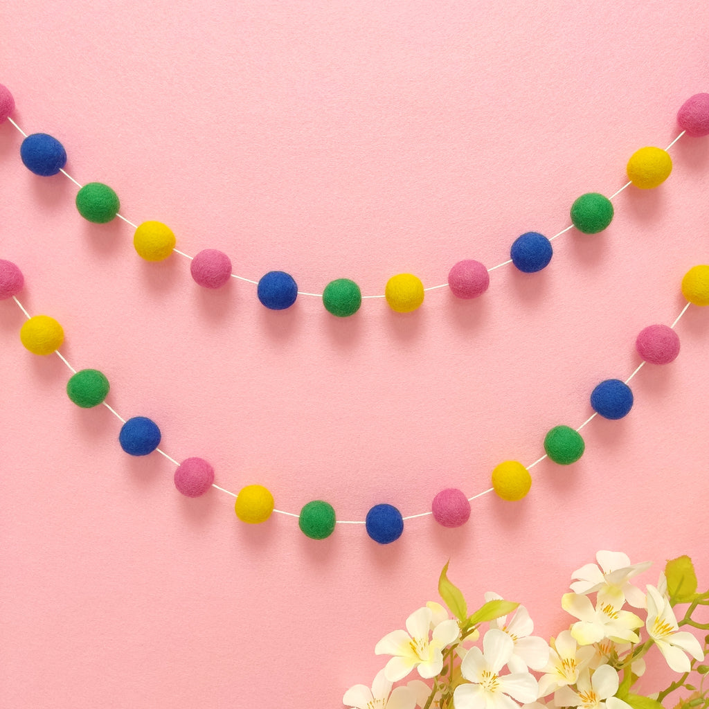 Guirlande de pompons feutrés confettis suspendue sur un mur, teintes bleu indigo, vert prairie, jaune soleil et rose bonbon