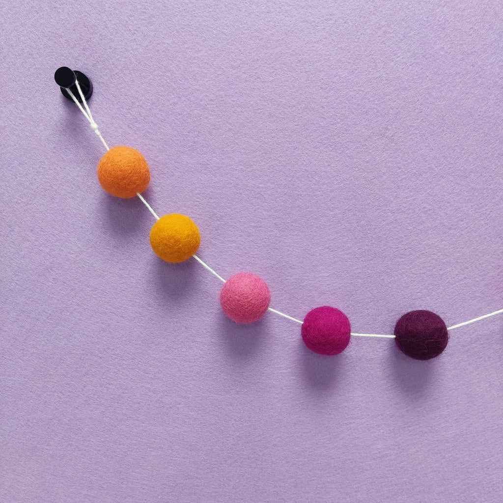 Zoom sur les 5 couleurs de la guirlande de pompons dahlia : orange marigold, orange carotte, rose bonbon, rose fuchsia, violet pourpre