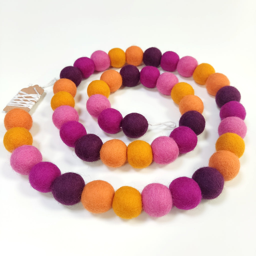 Guirlande en boule de laine feutrée dahlia rose, orange et pourpre disposée en spirale sur une table vue de dessus