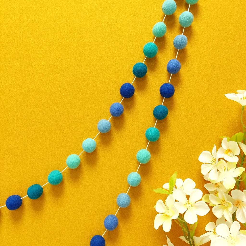 Guirlande de pompons feutrés aux teintes bleu indigo et bleu turquoise accrochée sur un mur