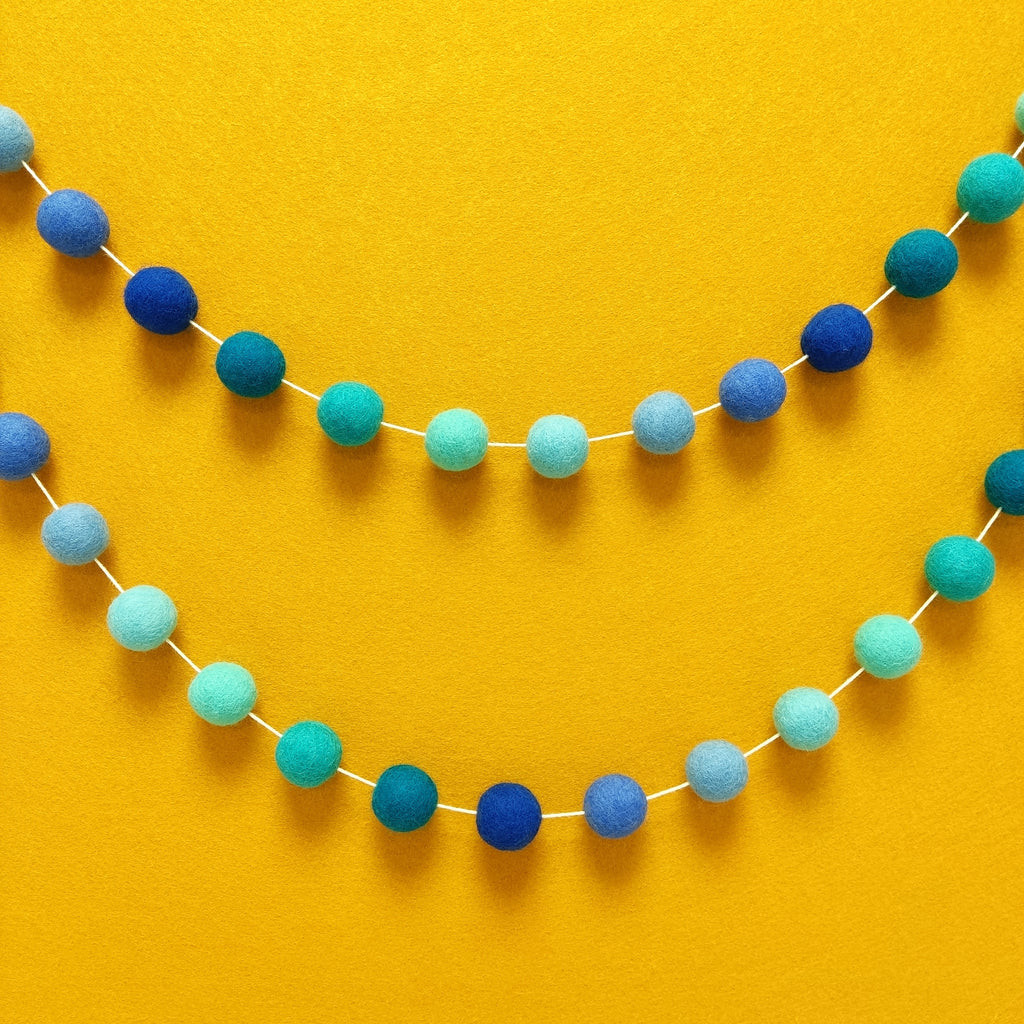 Guirlande de pompons en feutre de laine mérinos aux couleurs turquoise et nuances de bleu suspendue en arc de cercle sur un mur