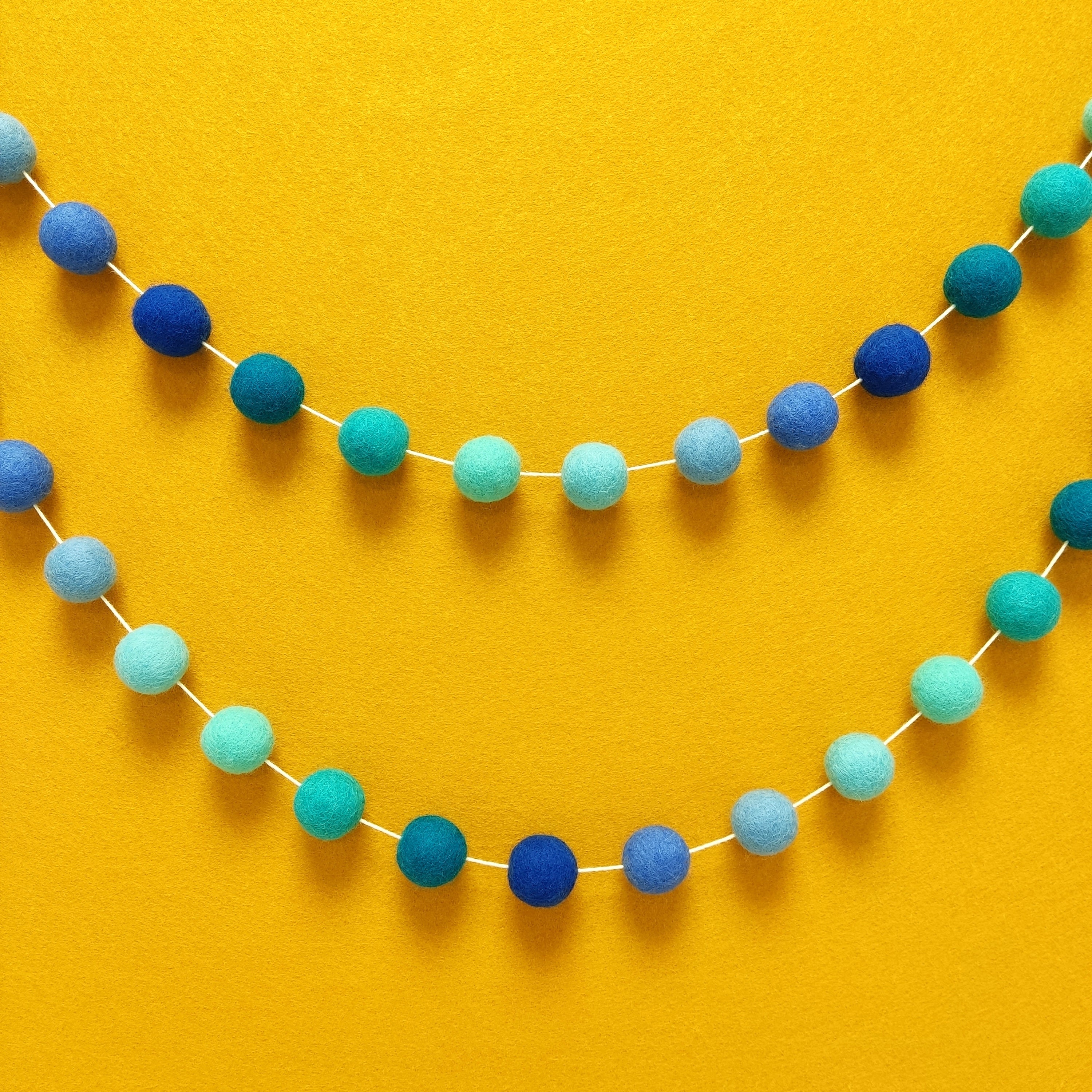 Guirlande de pompons en feutre de laine mérinos aux couleurs turquoise et nuances de bleu suspendue en arc de cercle sur un mur