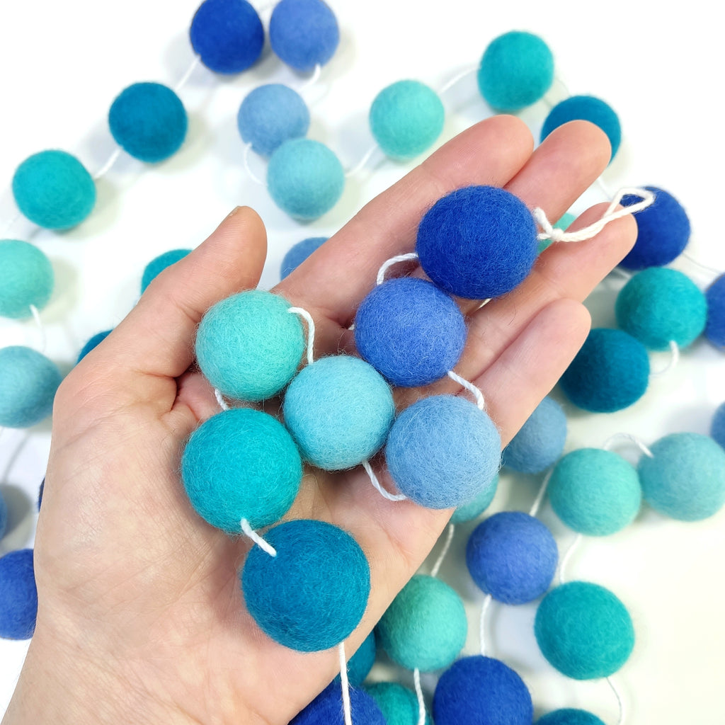 Zoom sur les pompons de feutre de 2,5 cm bleu turquoise, indigo, lagon, canard, lavande de la guirlande dégradé bleu dans une main
