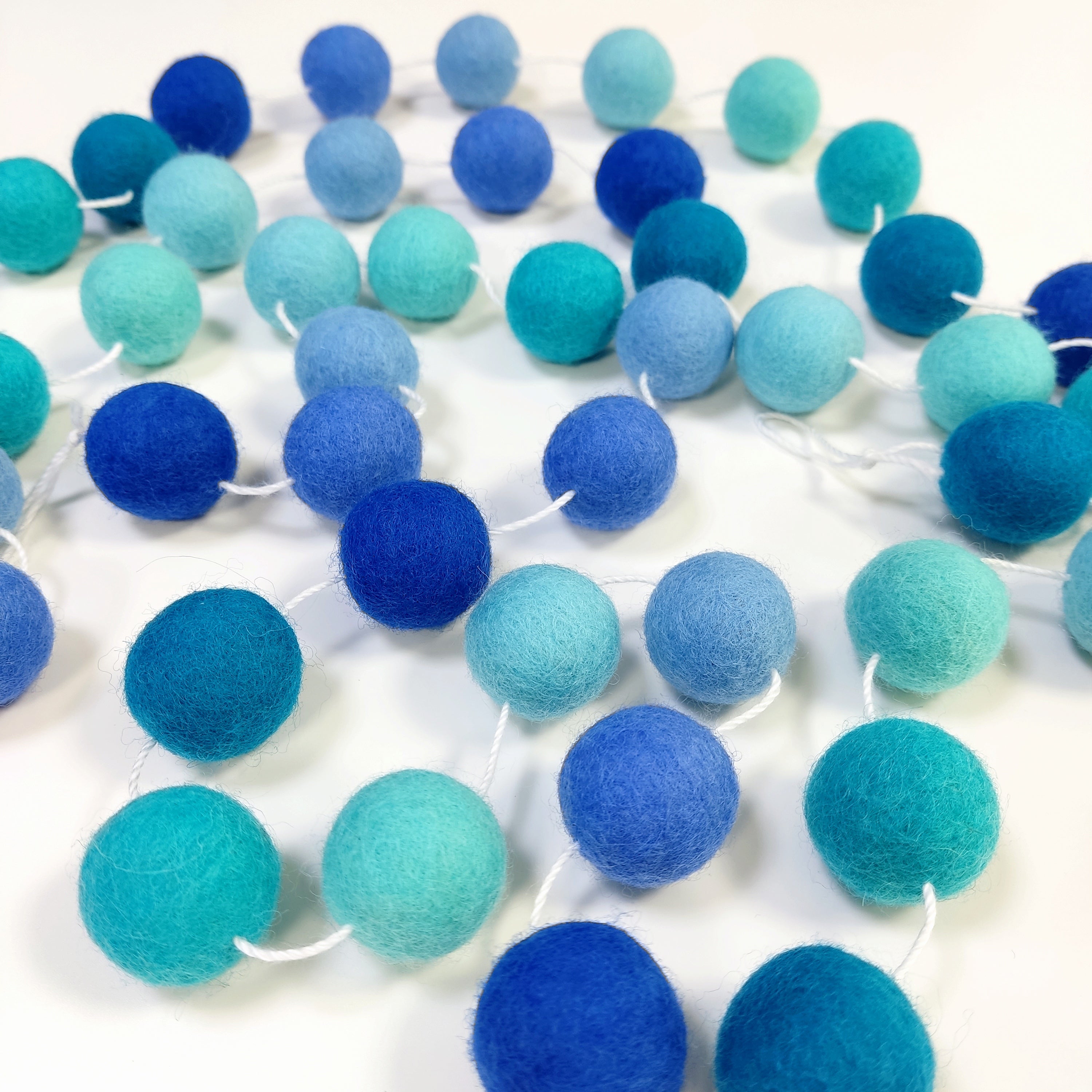 Guirlande de pompons en laine feutrée aux nuances bleu turquoise, ciel et indigo disposée sur une table avec les pompons espacés